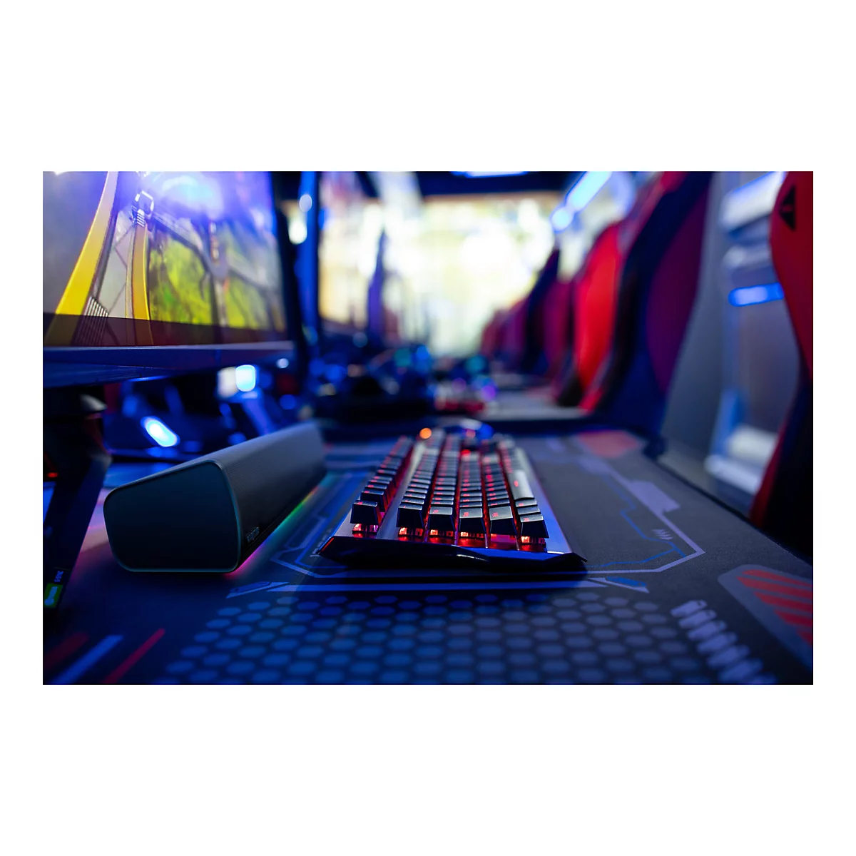 Eine Nahaufnahme einer beleuchteten Gaming-Tastatur, die sich auf einem Gaming-Schreibtisch befindet. Im Hintergrund sind Monitore und eine Gaming-Umgebung zu sehen.