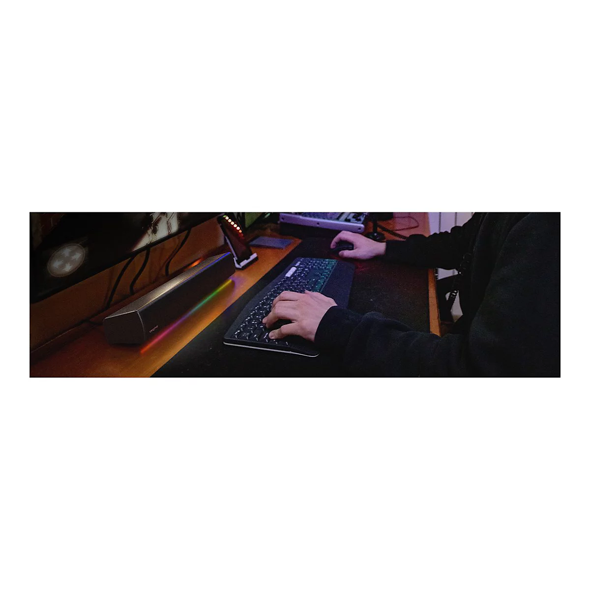 Person mit Tastatur, Maus, Monitor und Soundbar. Soundbar mit farbiger Beleuchtung.