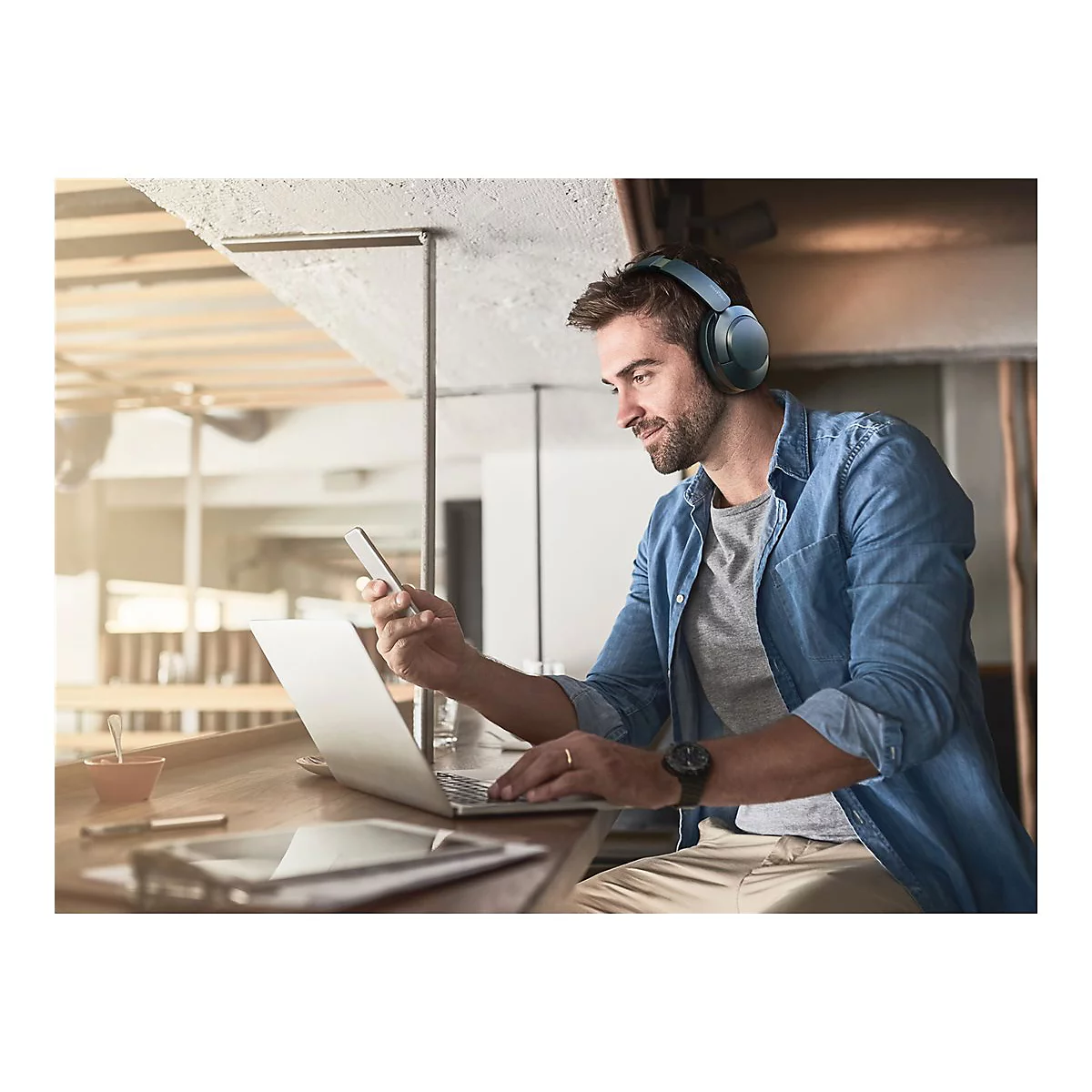 Mann mit KopfhÃ¶rern am Laptop, Smartphone in der Hand. Er sitzt in einem CafÃ©.