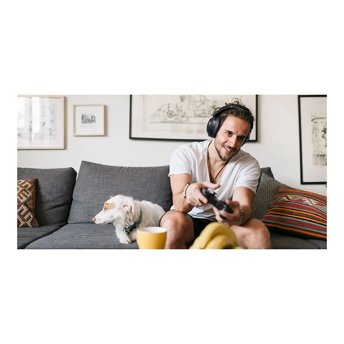 Ein Mann spielt mit KopfhÃ¶rern und Controller auf einer Couch. Ein Hund und eine Tasse sind auch zu sehen.