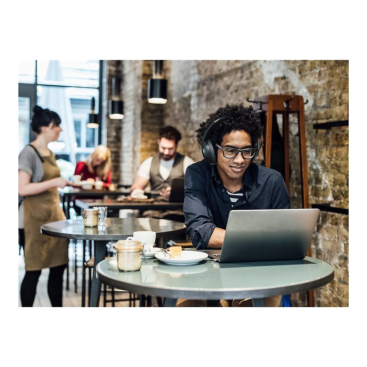 Mann mit Brille und KopfhÃ¶rern, der an einem Laptop in einem CafÃ© sitzt, lÃ¤chelt.