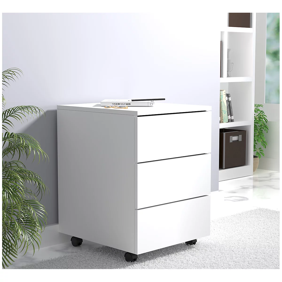 White three-drawer filing cabinet on wheels. Pens and notes on top. Background shelf and houseplant.