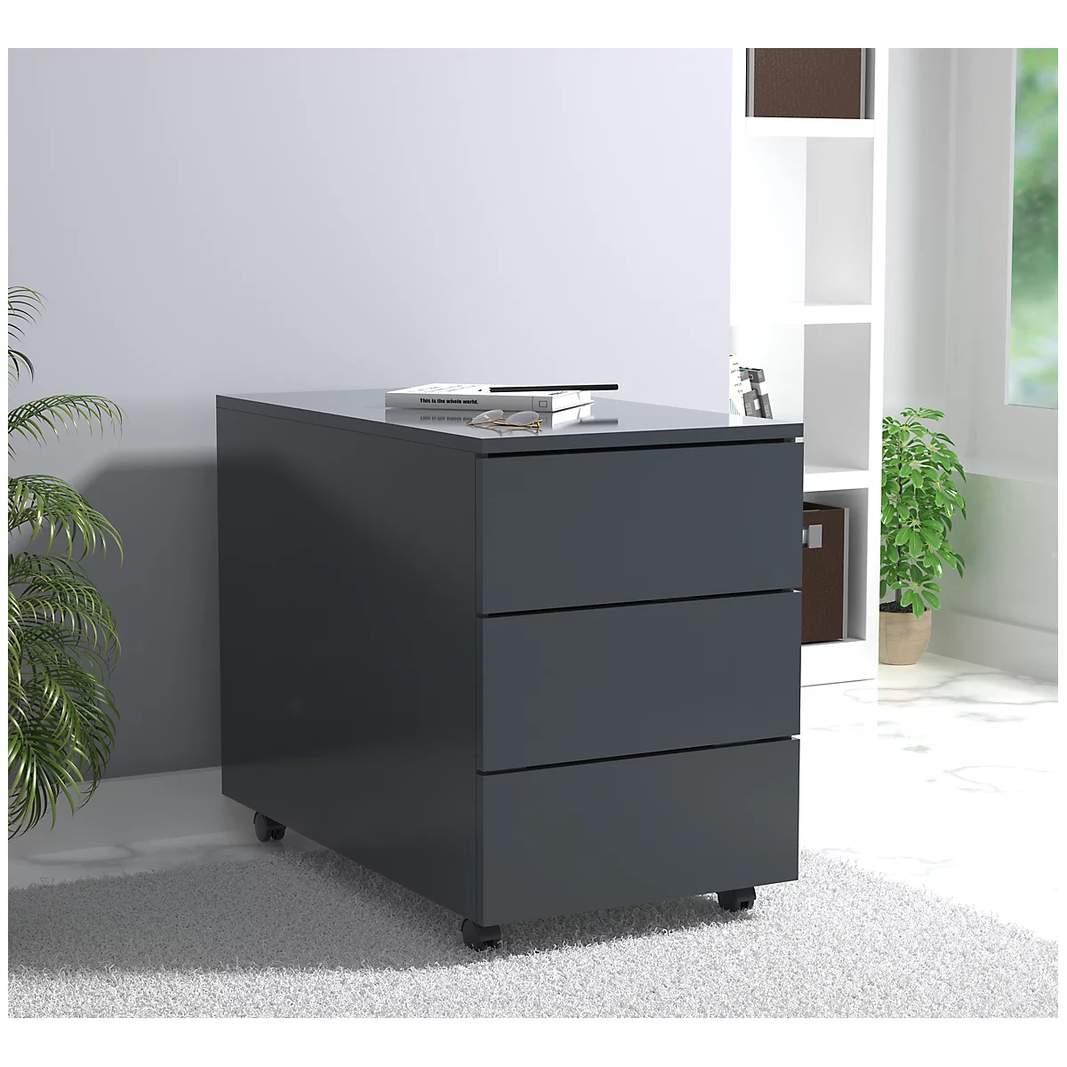 Dark gray mobile pedestal with three drawers. A book and glasses lie on top. In the background: shelf, plant.