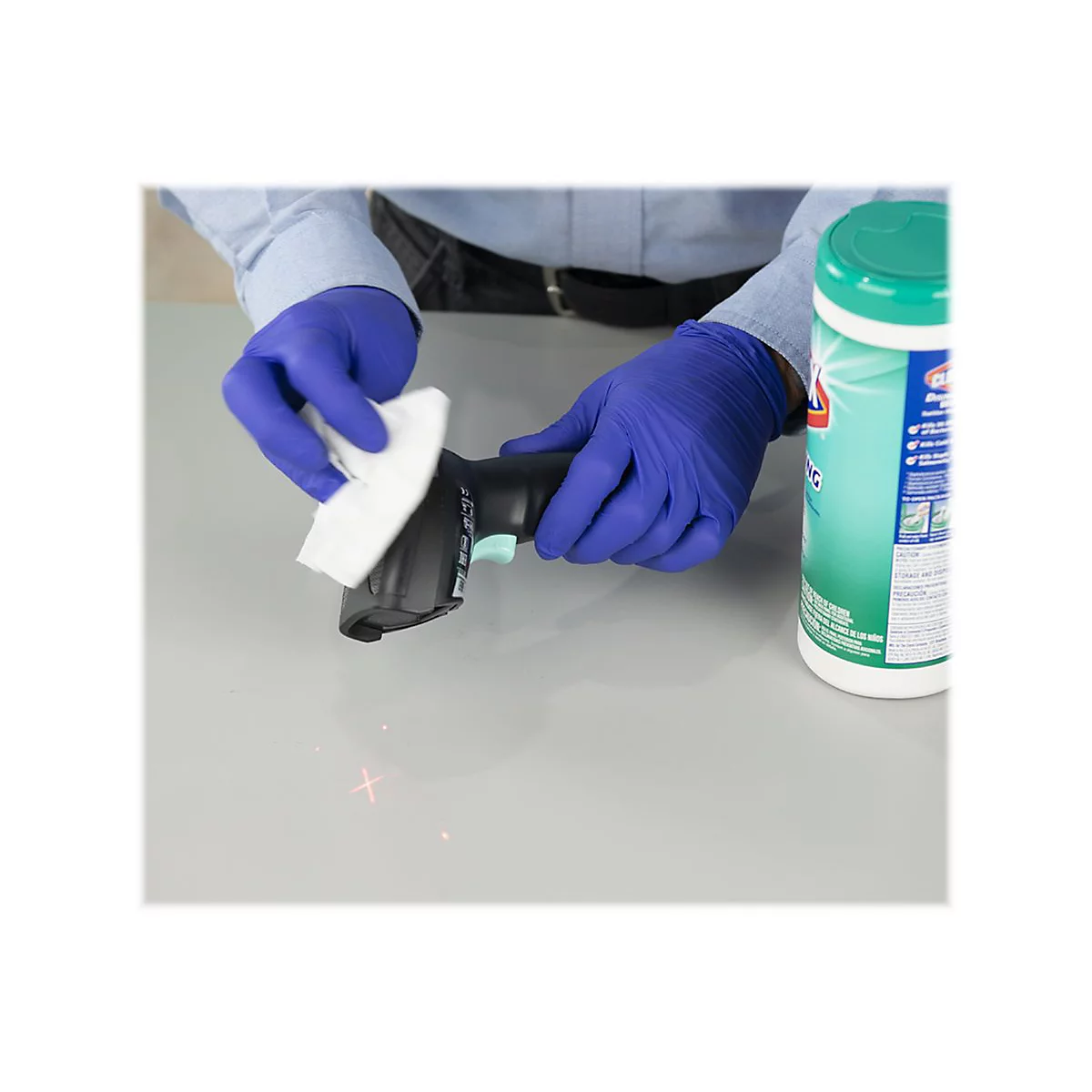 Person reinigt Barcode-Scanner mit einem Wischtuch, trÃ¤gt blaue Handschuhe, daneben DesinfektionstÃ¼cher.