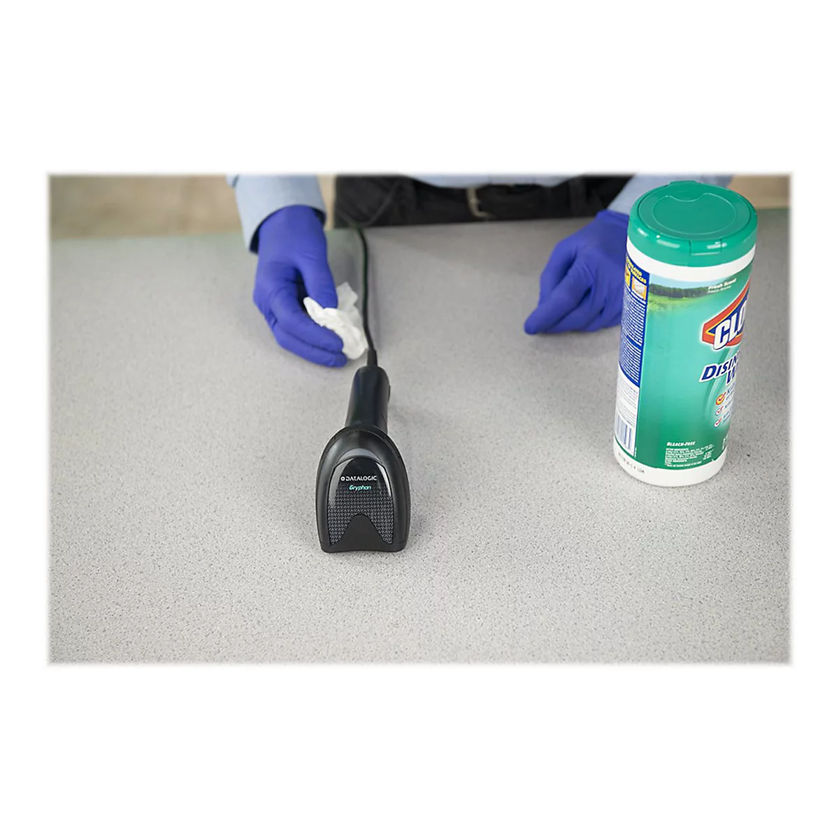 Person mit Handschuhen, hält ein Reinigungstuch über einen Barcode-Scanner, daneben eine Desinfektionstuch-Dose.
