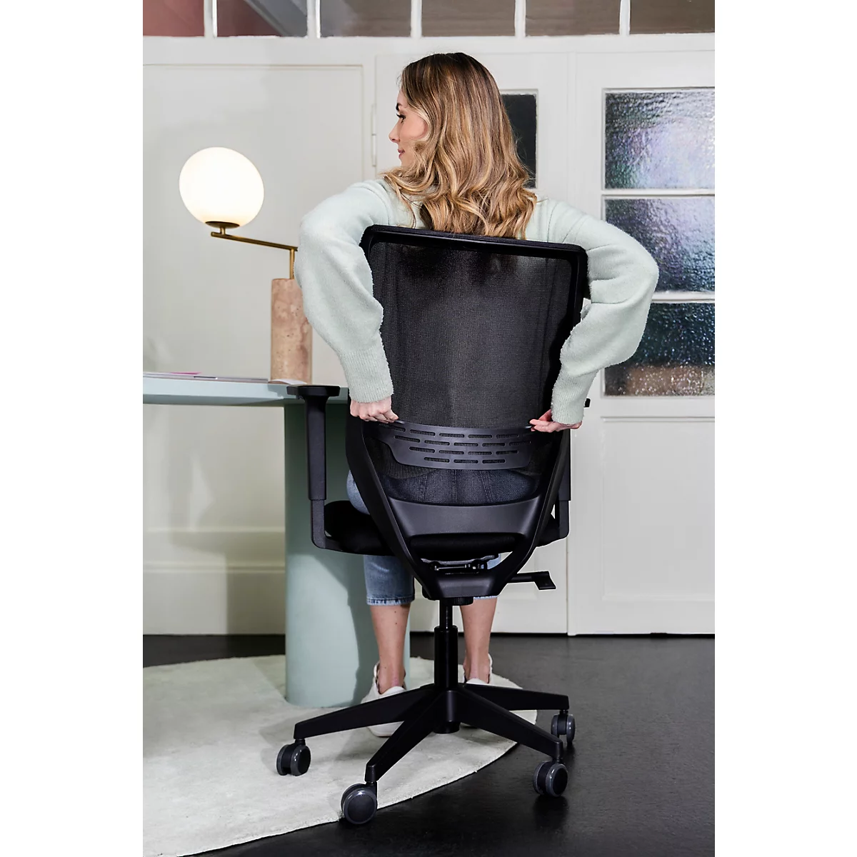 Eine Frau sitzt auf einem schwarzen Bürostuhl, ihre Hände am Stuhl. Der Hintergrund ist unscharf, mit einem Schreibtisch und einer Lampe.