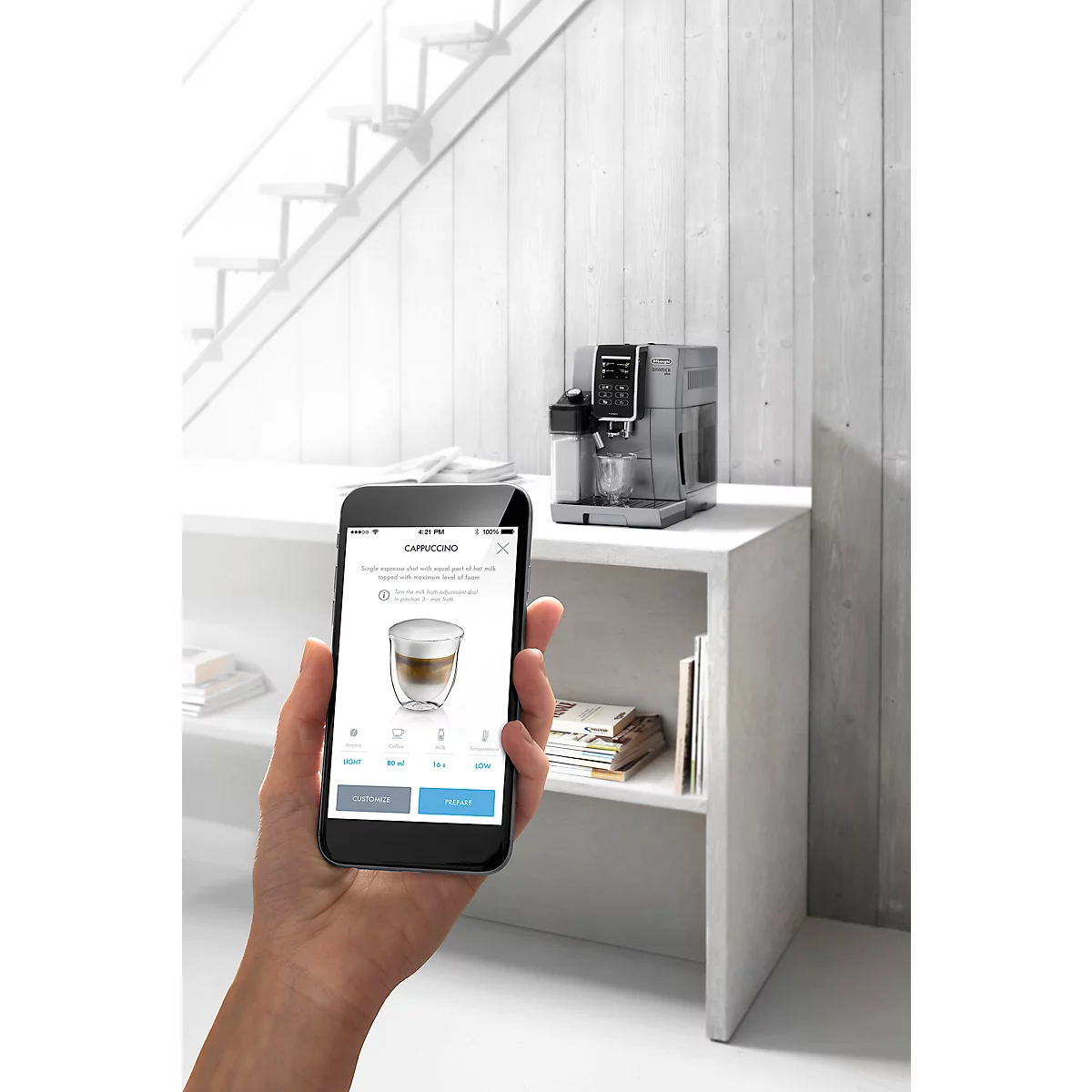 Hand hält Smartphone mit Cappuccino-Auswahl. Kaffeemaschine und Regalbrett im Hintergrund.