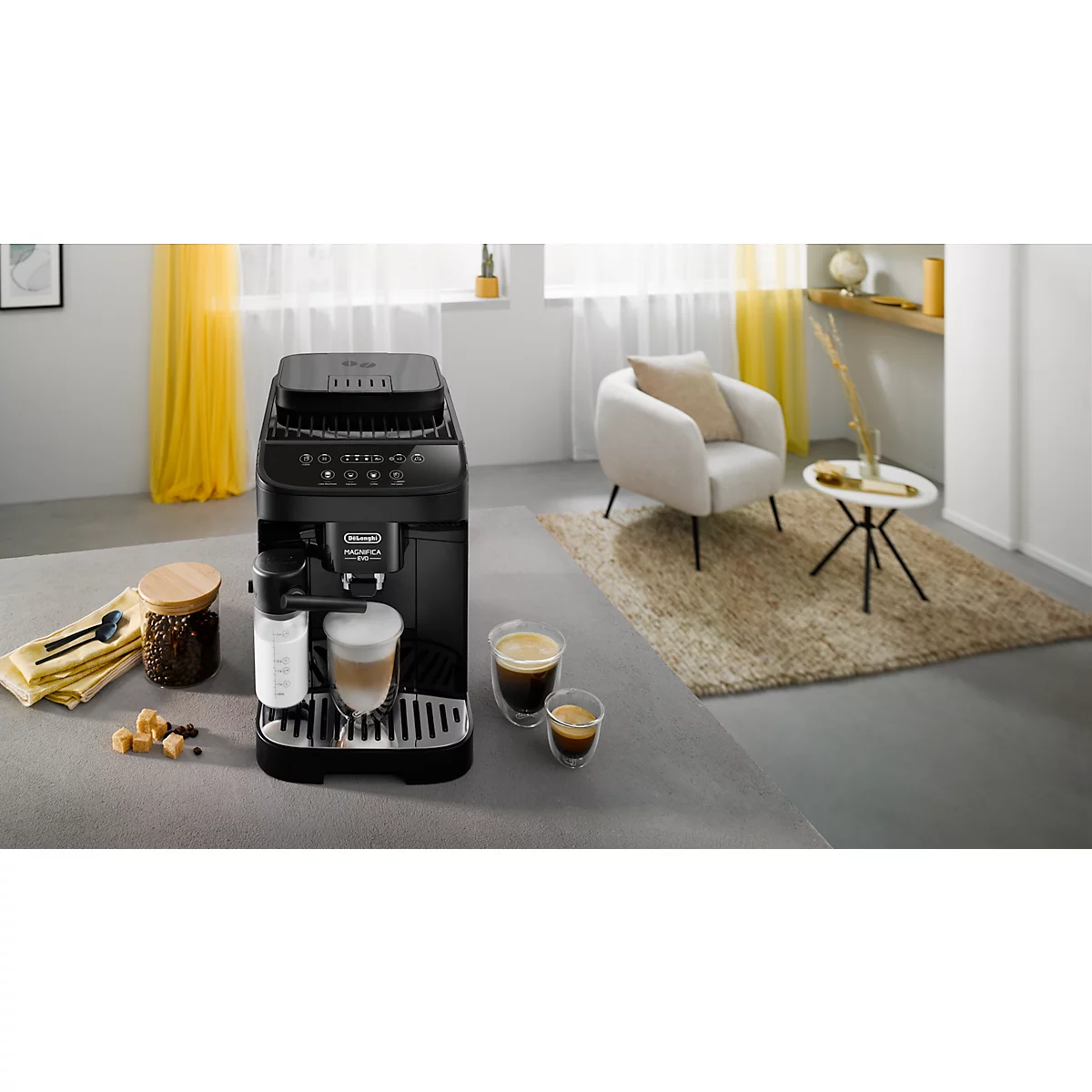 Schwarze Kaffeemaschine in einem Wohnzimmer, daneben eine Tasse Kaffee und ein gemütlicher Sessel. Helle Atmosphäre.
