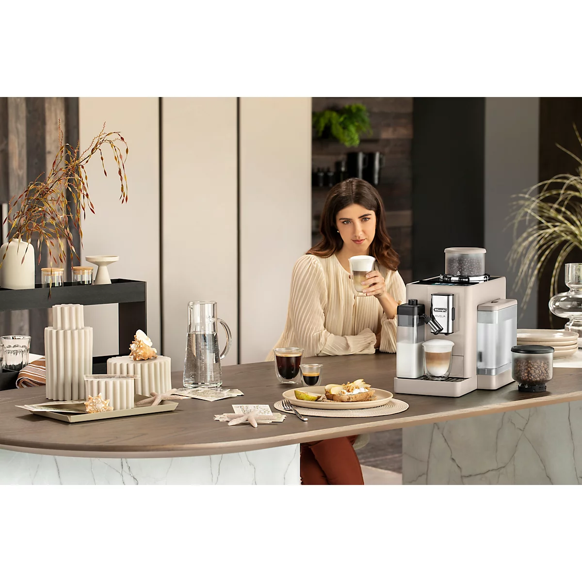 Eine Frau mit einem Cappuccino sitzt an der Theke neben einer Kaffeemaschine. Auf dem Tisch stehen auch Kaffee, Gebäck und Dekoration.