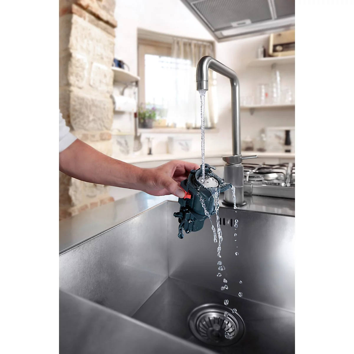 Een persoon spoelt een donker apparaat af onder stromend water in een gootsteen. Op de achtergrond is een keuken te zien.