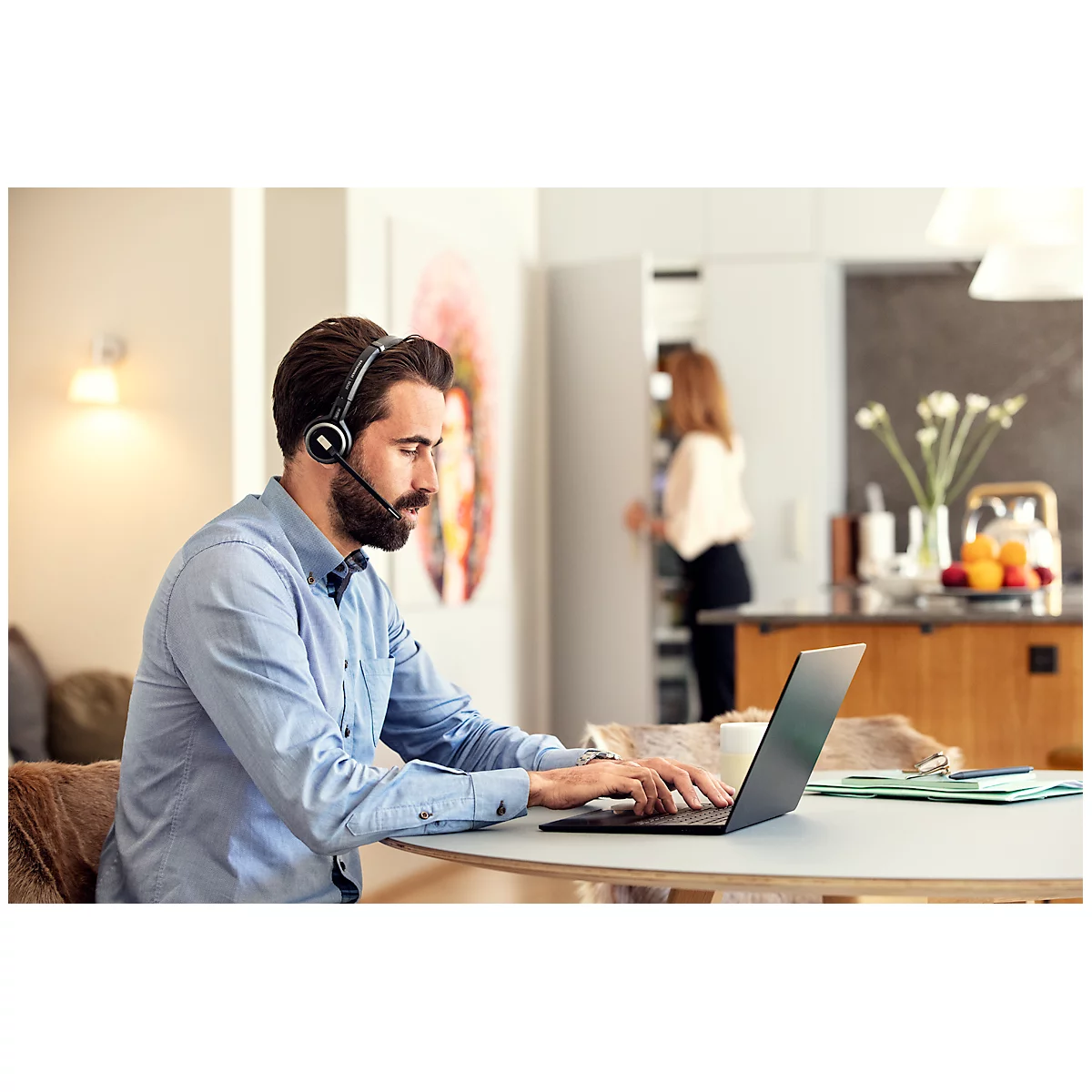 A man with a headset types on a laptop at a table. A woman is in the background in the kitchen.