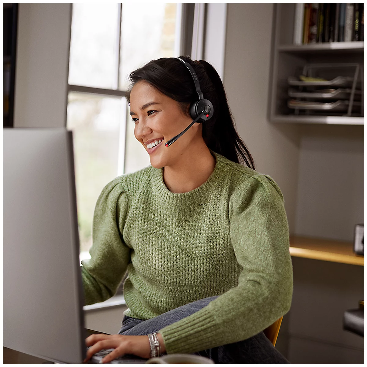 Smiling woman with a headset in front of a PC. She is sitting at a desk with a window in the background.