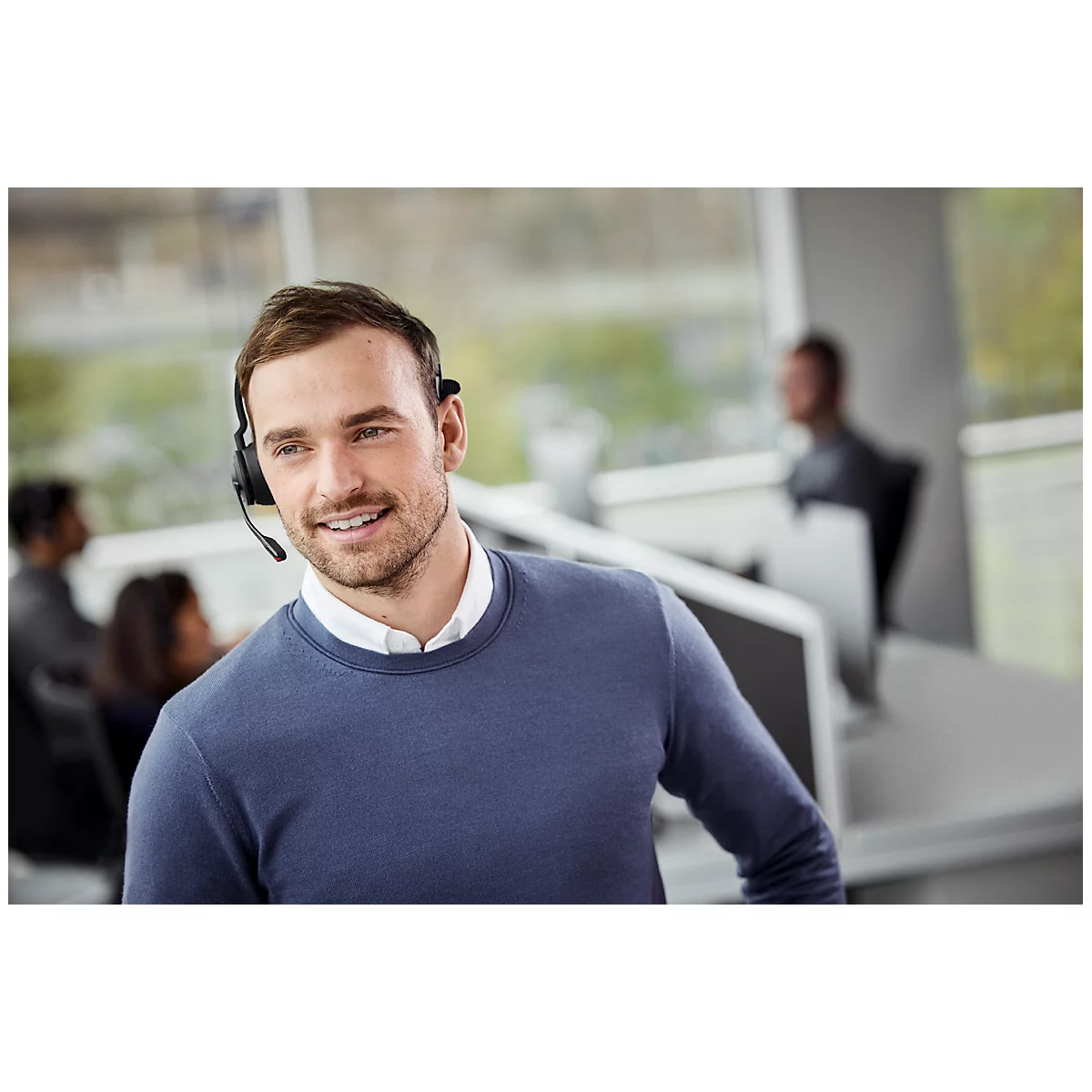 A smiling man wearing a headset in an office. Other people and screens are visible in the background.