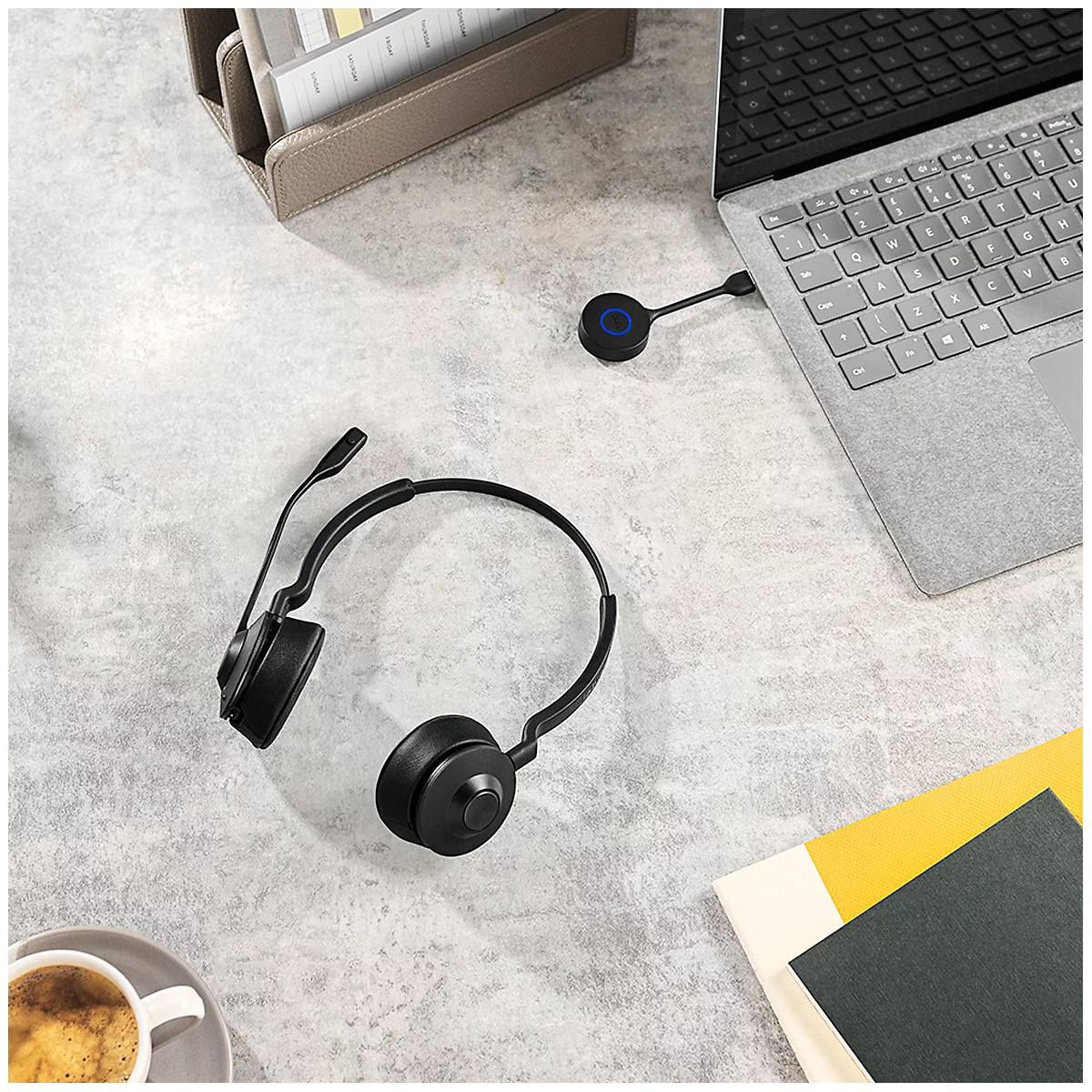 Black headset lies next to laptop, coffee cup and notebooks on a light desk.