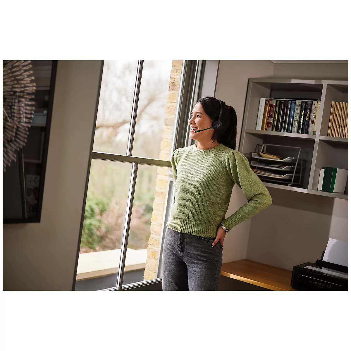 Woman with headset looks out of a window in an office. She smiles relaxed, arm on hip.