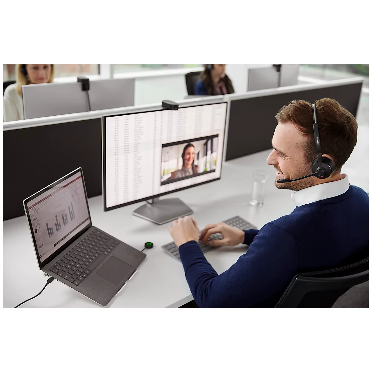 A man wearing a headset at a desk, in a video call, a woman can be seen on the monitor, other people in the background.