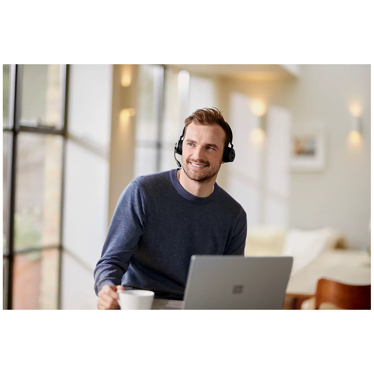 A man wearing a headset smiles while working on a laptop, holding a mug.