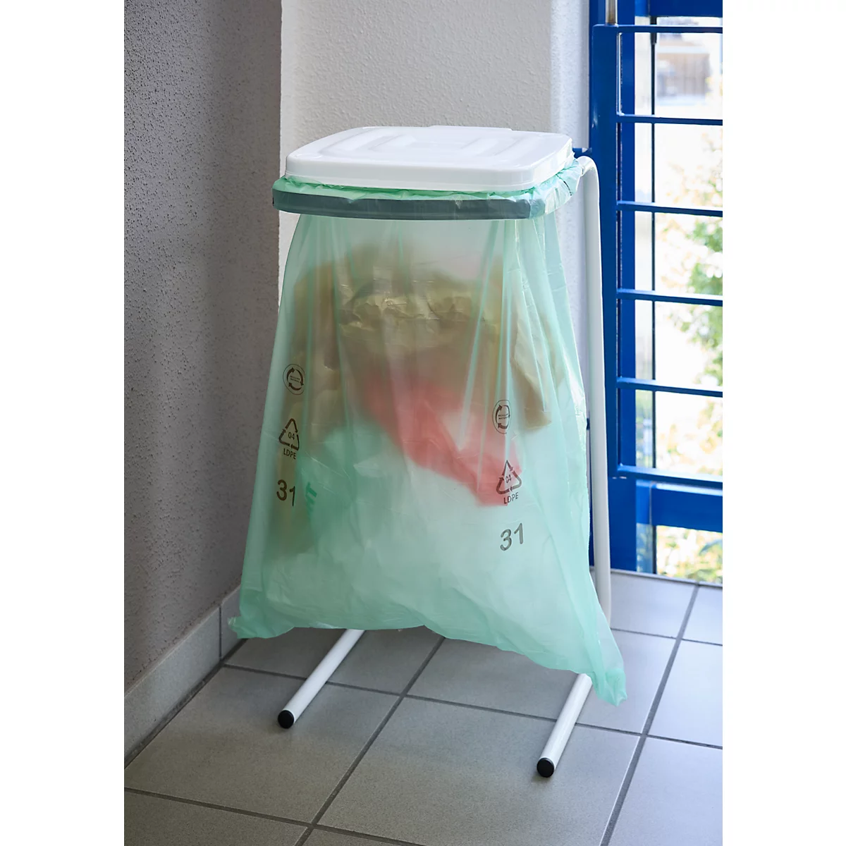 Ein grüner Müllsack in einem weißen Gestell mit Deckel. Im Hintergrund ist eine blaue Fensterstruktur zu sehen.