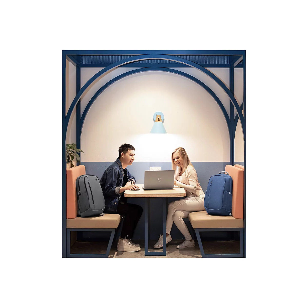 Zwei Personen sitzen an einem Tisch und schauen auf einen Laptop, vor einem blauen Bogen, auf den Sitzen stehen RucksÃ¤cke.
