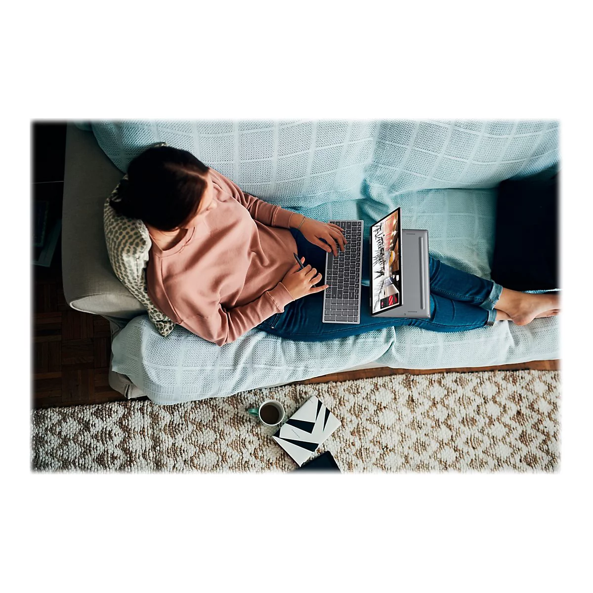 Junge Frau entspannt auf Couch, tippt auf Laptop. Kaffeetasse, Notizen auf Teppich. Angenehme Arbeitsatmosphäre.