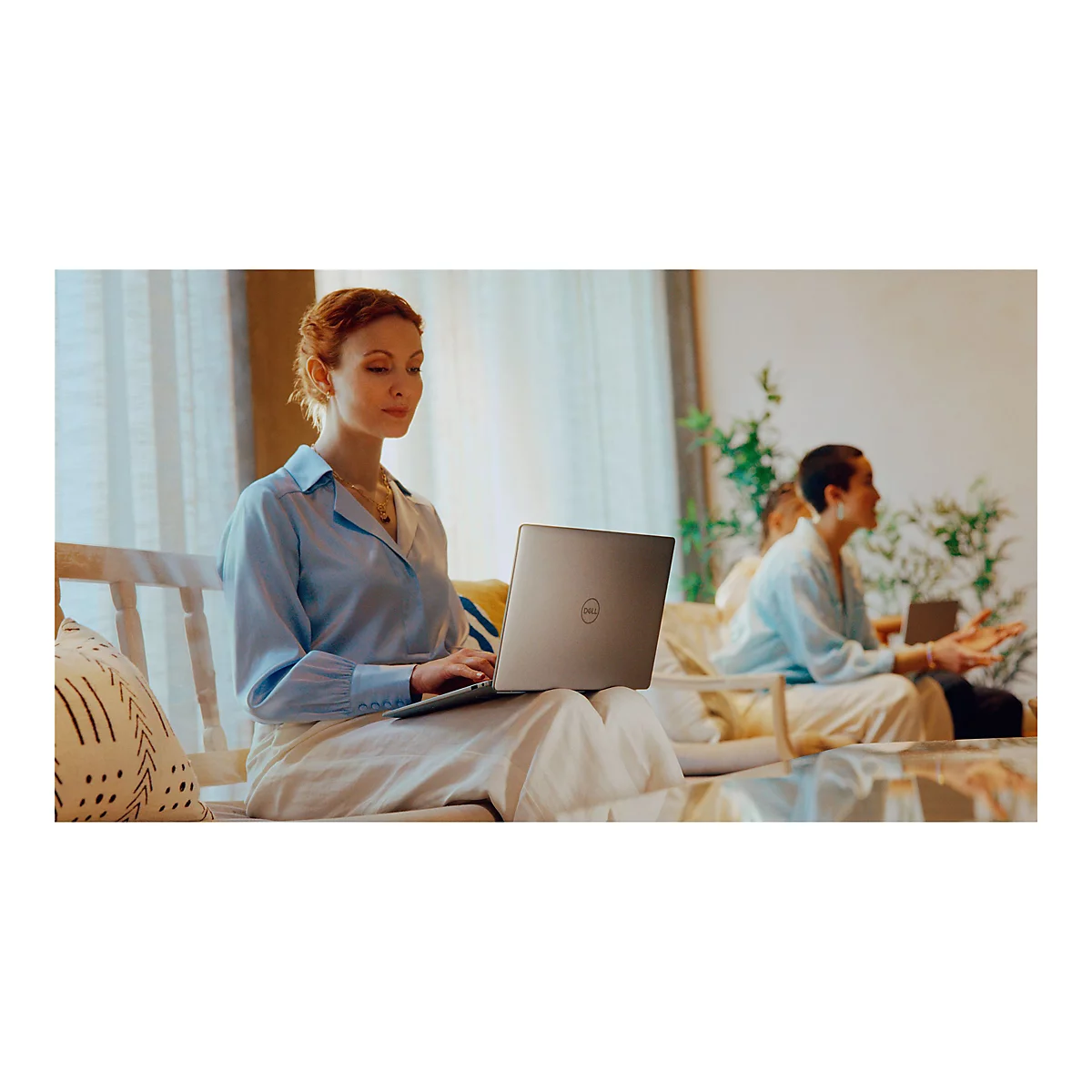 Eine Frau mit roten Haaren sitzt auf einem Sofa und arbeitet an einem silbernen Laptop. Eine weitere Person sitzt im Hintergrund.