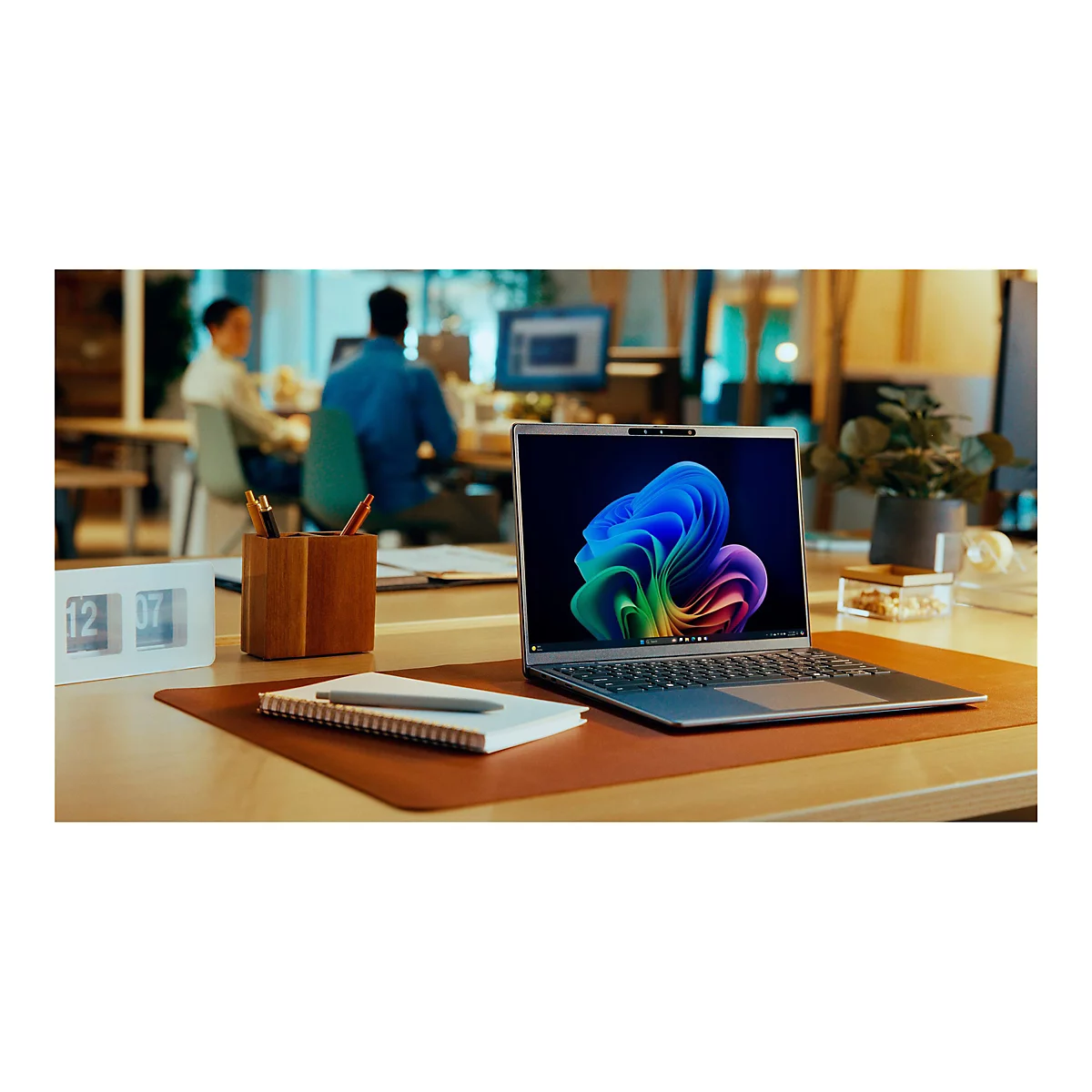 Ein Laptop mit Windows-Hintergrund steht auf einem Schreibtisch, im Hintergrund arbeiten zwei Personen.