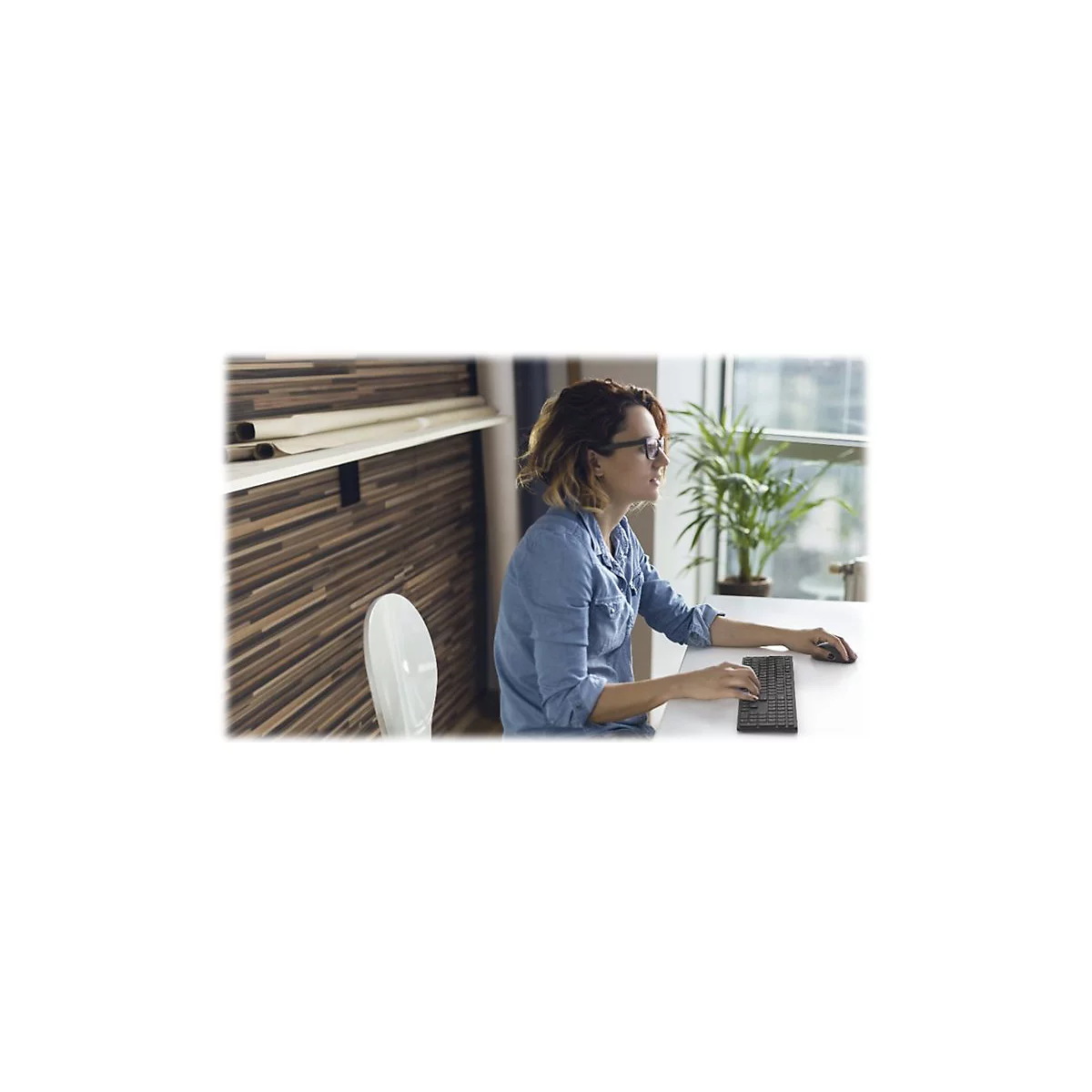 Eine Frau sitzt an einem Tisch und arbeitet am Computer. Sie trägt eine Jeansjacke und Brille.
