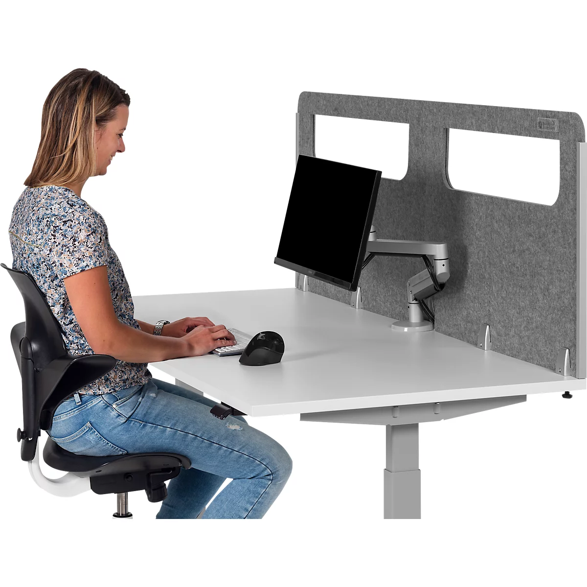 Woman sitting at a height-adjustable desk with a monitor, typing on a keyboard. Gray wall, black office chair, light wood.