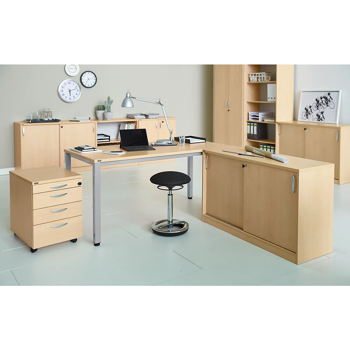 A modern office with a desk, mobile pedestal, chair, lamps, filing cabinets, and clocks on the wall.