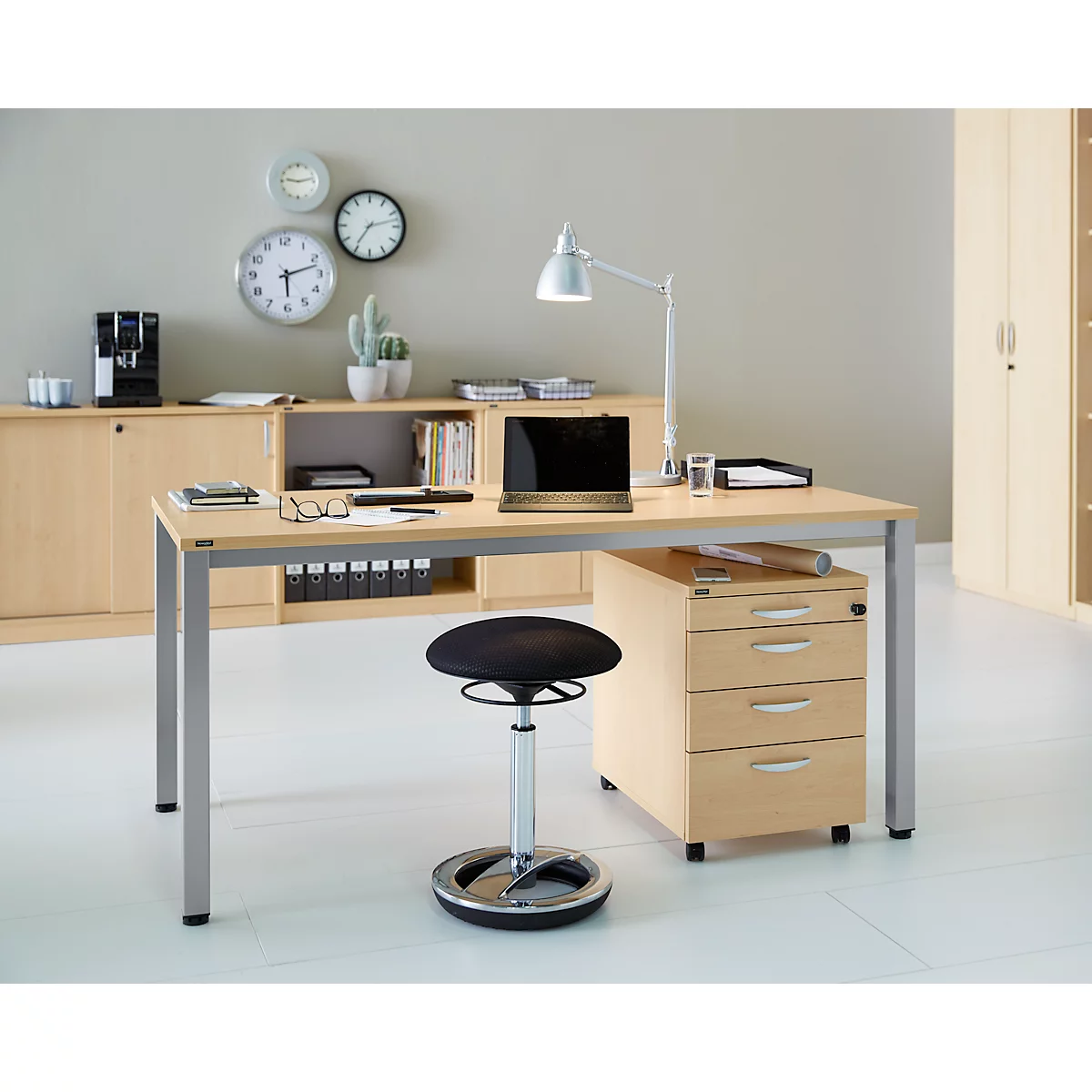 Office setup with desk, chair, lamp, laptop, and cabinets. Clocks and a plant are visible on the wall.