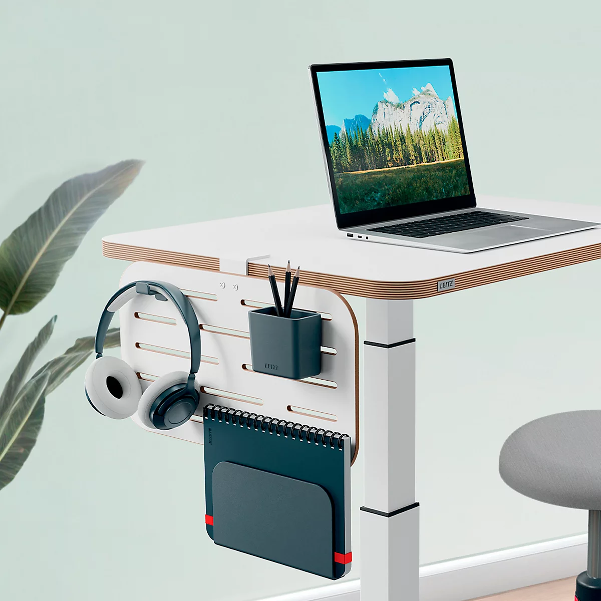 Desk with laptop, headphones, and notebook. Background: light green. A mountain landscape is visible on the laptop.