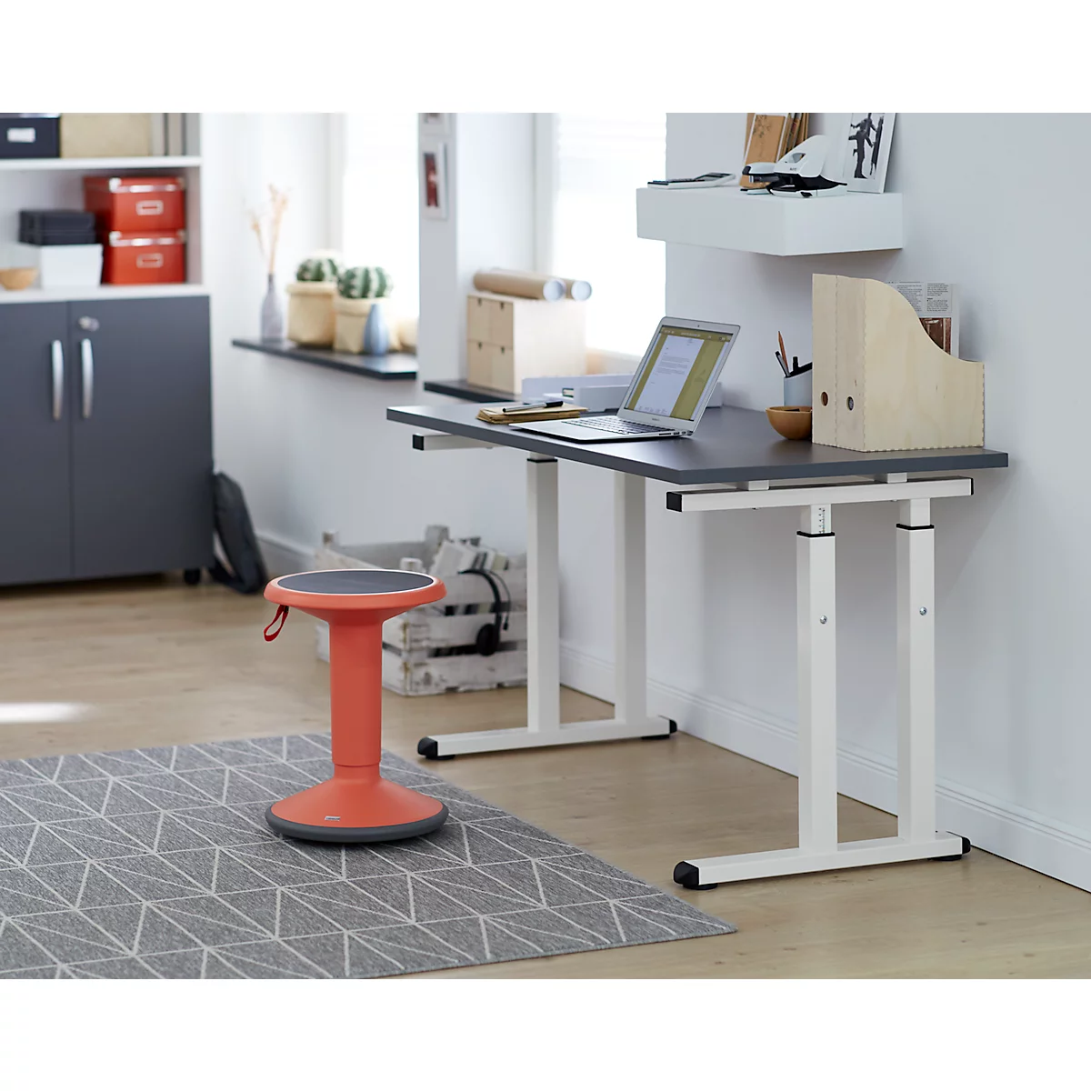 Desk with laptop and red stool, stands in front of window with light walls, on gray carpet.
