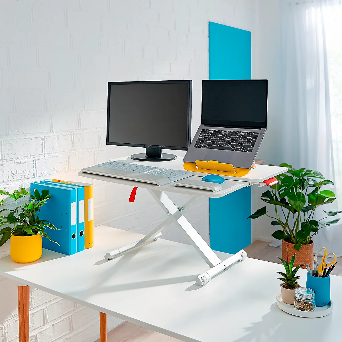 A white, height-adjustable desk with computer, laptop and plants in front of a white brick wall.