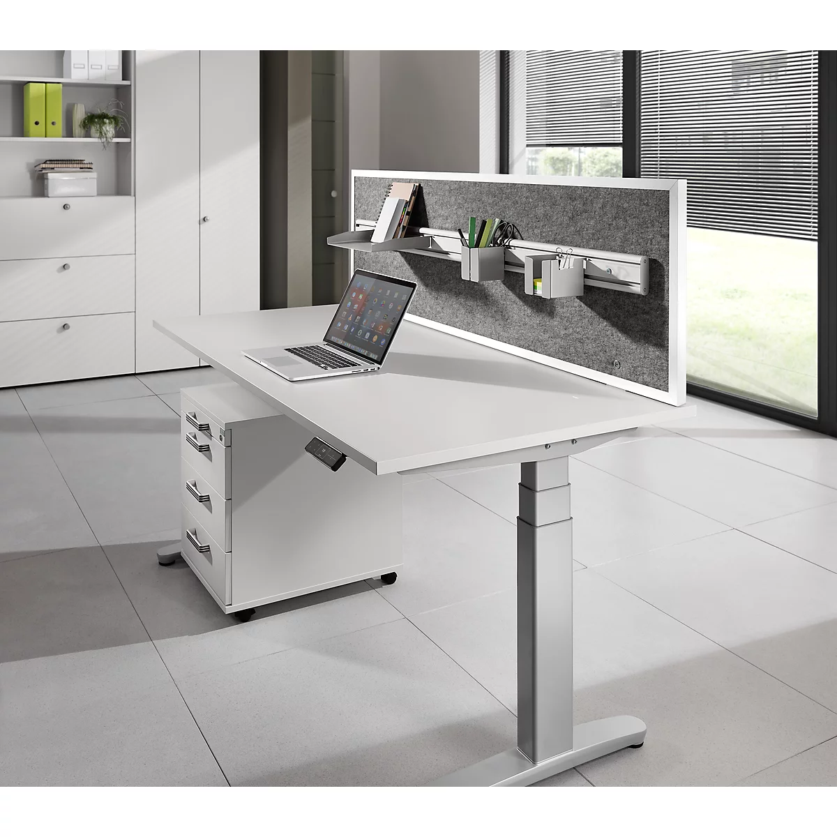 White height-adjustable desk with laptop, gray felt board, pen holders, and cabinet. In the background: white cabinet.