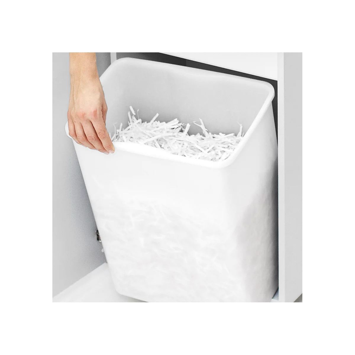 Une main tient une corbeille à papier blanche remplie de papier déchiqueté. La corbeille à papier est intégrée dans une armoire.