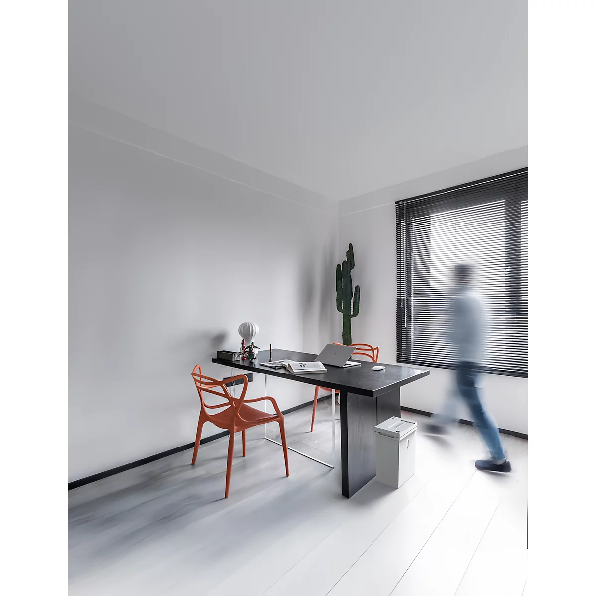 Bureau avec bureau, chaises et cactus. Une personne floue passe. Aspect bois clair et fenêtre avec stores.