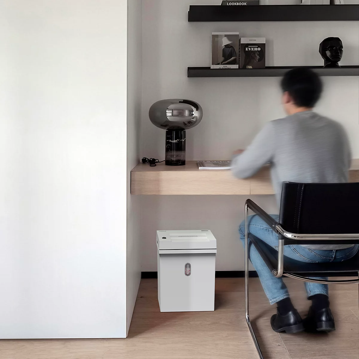 Un homme est assis à un bureau minimaliste. Il y a une lampe sur le bureau. A côté du bureau se trouve un destructeur de documents.