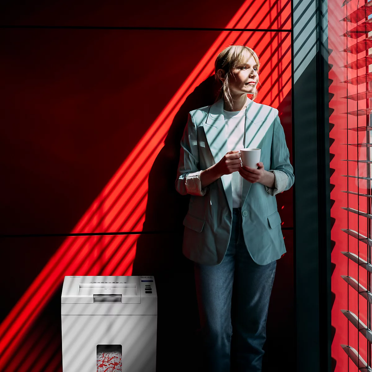 Femme avec café à la fenêtre, la lumière du soleil projette des ombres sur le mur rouge. Destructeur de documents à gauche.