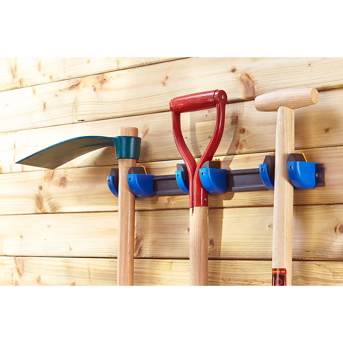 Three garden tools, a hoe, a spade and a shovel, are hanging on a wooden panel wall.