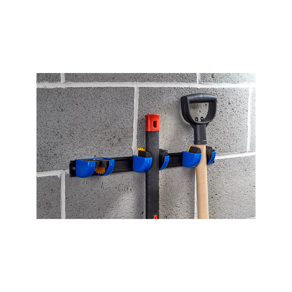 Tool holder with shovel and broom handle on a concrete wall.