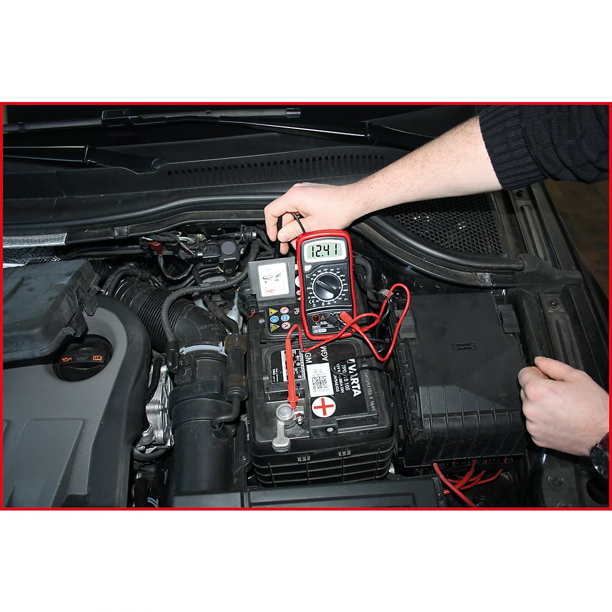 Ein rotes Multimeter misst die Spannung an einer Autobatterie. Das Display zeigt 12,41 V an. Das Bild zeigt den Motorraum eines Autos.