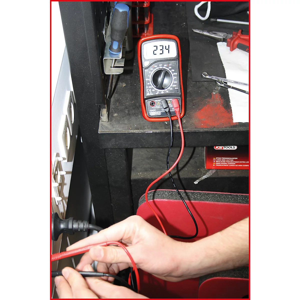 Ein Multimeter zeigt 234 an. Rote und schwarze Messleitungen sind angeschlossen, Hände halten weitere Leitungen. Werkbank.