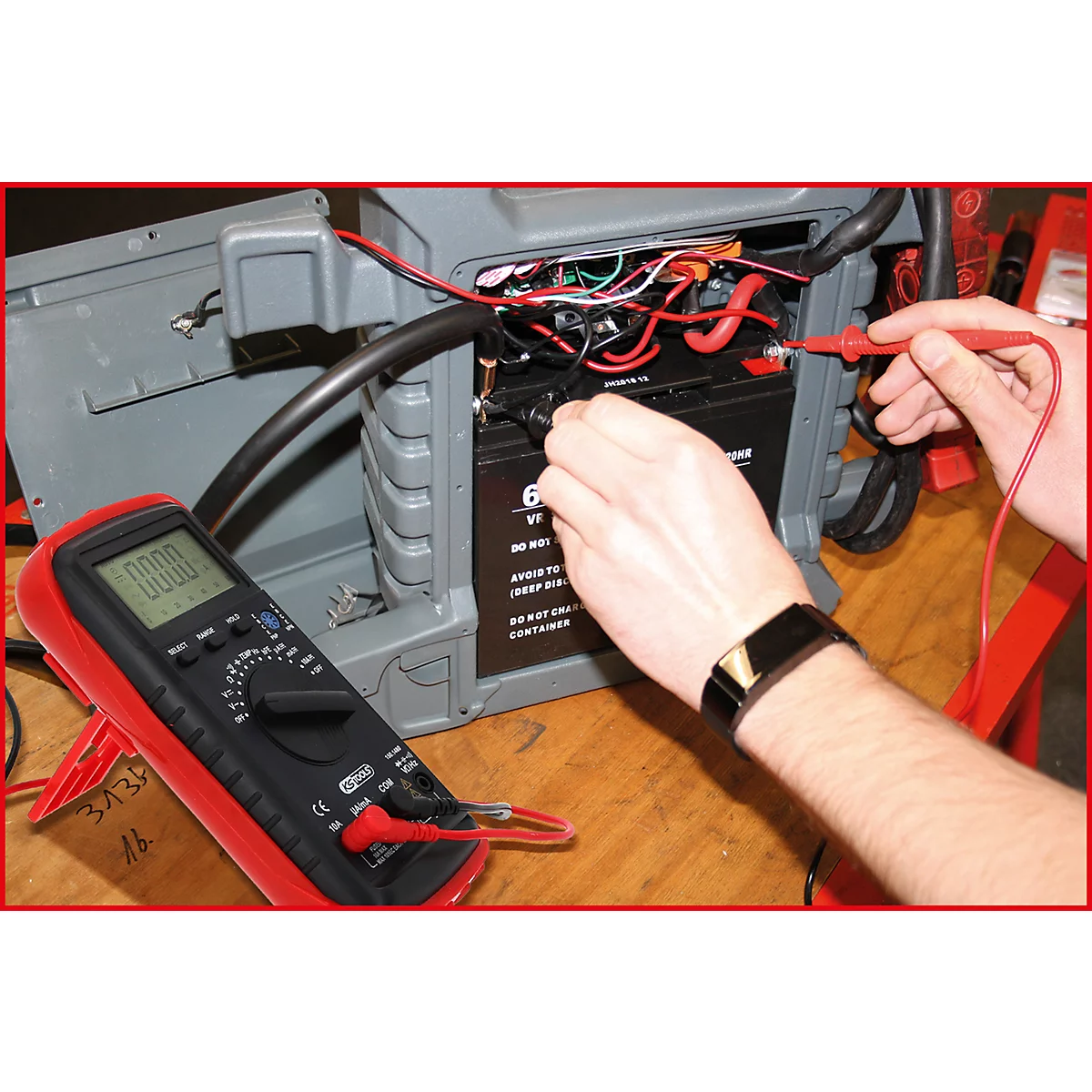 Hände prüfen mit einem Multimeter eine Batterie in einem grauen Gehäuse. Auf dem Multimeter steht 0.000.