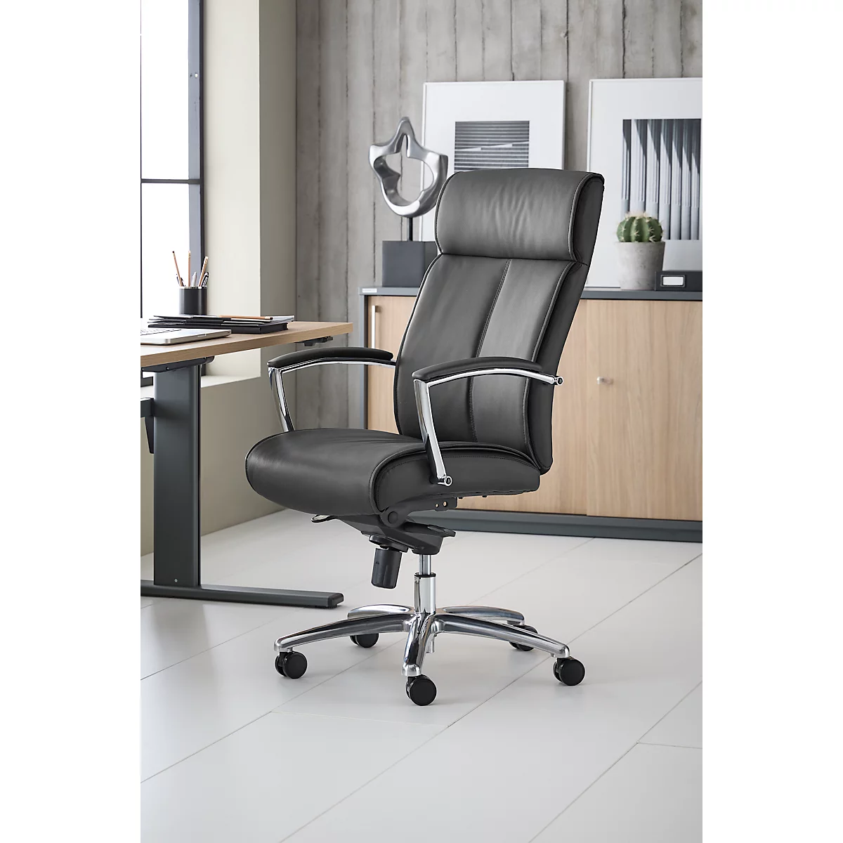 Zwarte bureaustoel, leer, chromen poten, tegen kantoorachtergrond.