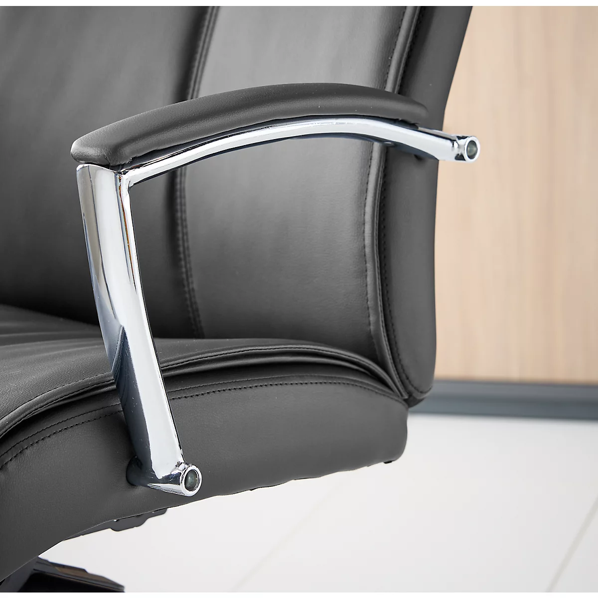 Detail van een zwarte bureaustoel, armleuning van leer en metaal. De stoel staat voor lichtbruin hout en een witte vloer.