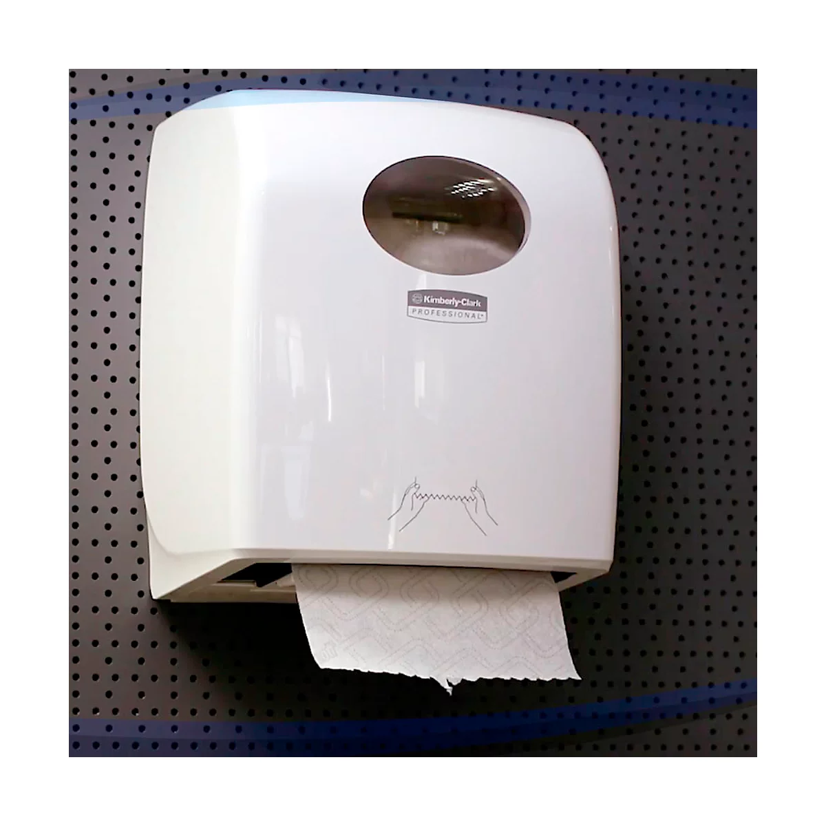 Distributeur d'essuie-mains en papier blanc sur tôle perforée grise. Fenêtre de visualisation ronde en haut, sortie de papier en bas. Inscription : Kimberly Clark Professional.