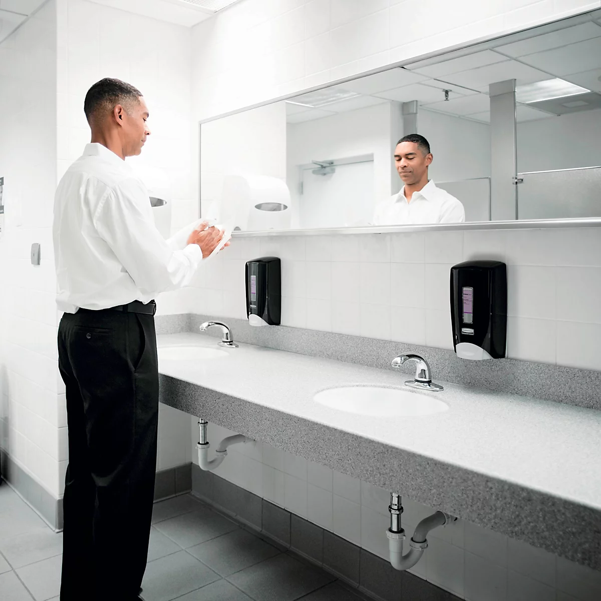 Un homme en chemise blanche se sèche les mains dans des toilettes publiques. On peut voir son reflet.