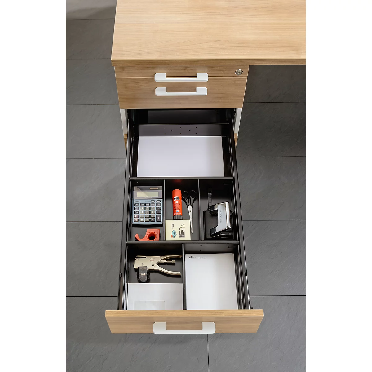 Open desk drawer with office supplies such as calculator, stapler, scissors, paper, and tape.