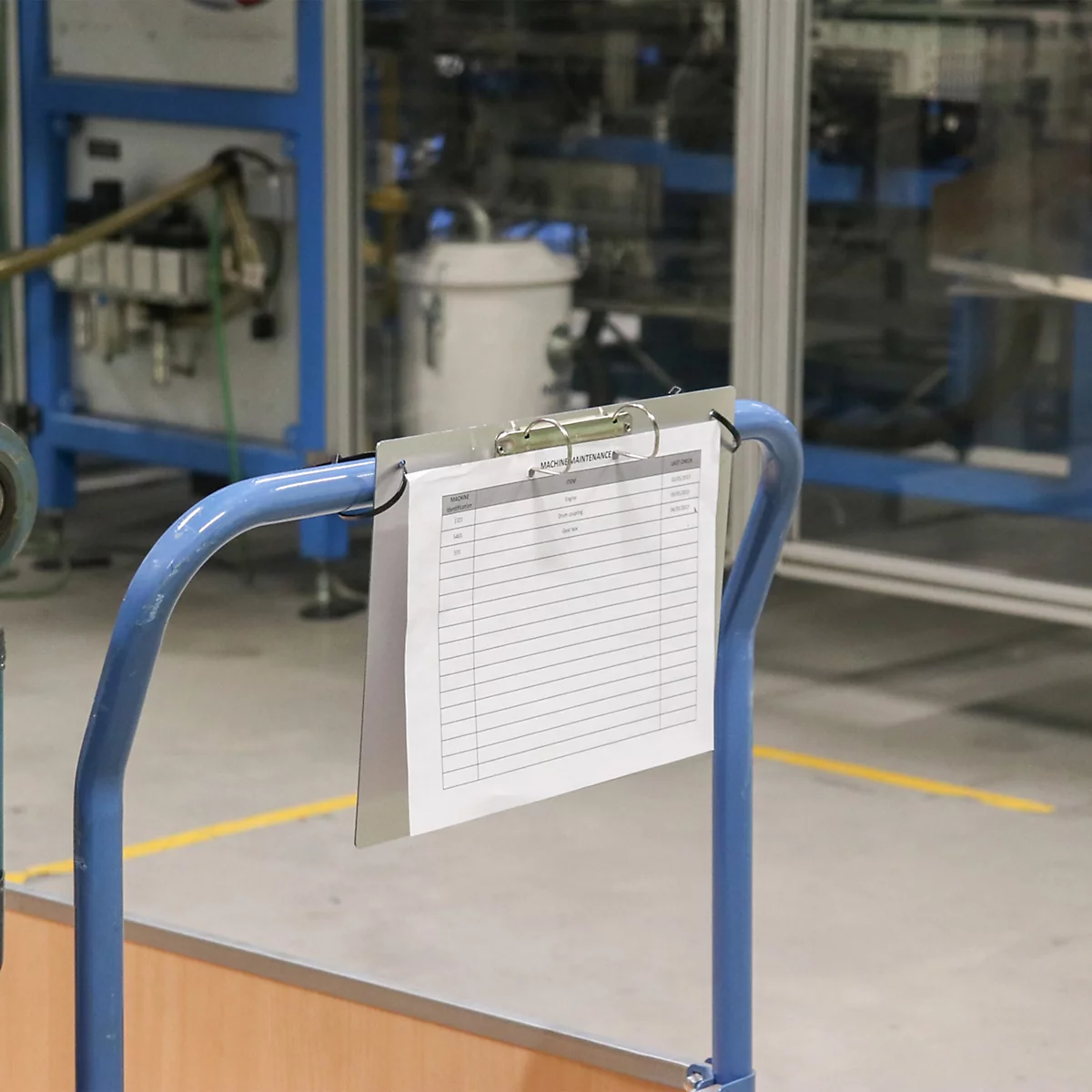 Klemmbrett mit Formular hängt an blauem Metallgestell in einer Fabrikhalle.