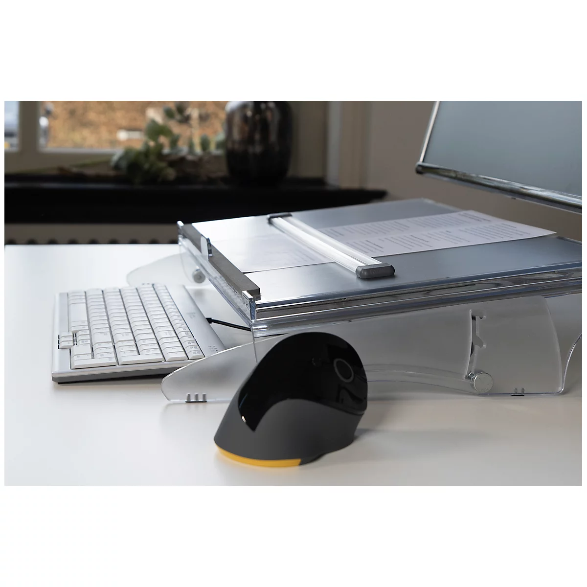 A white desk displays a keyboard, mouse, and paper. Windows and a vase are visible in the background.